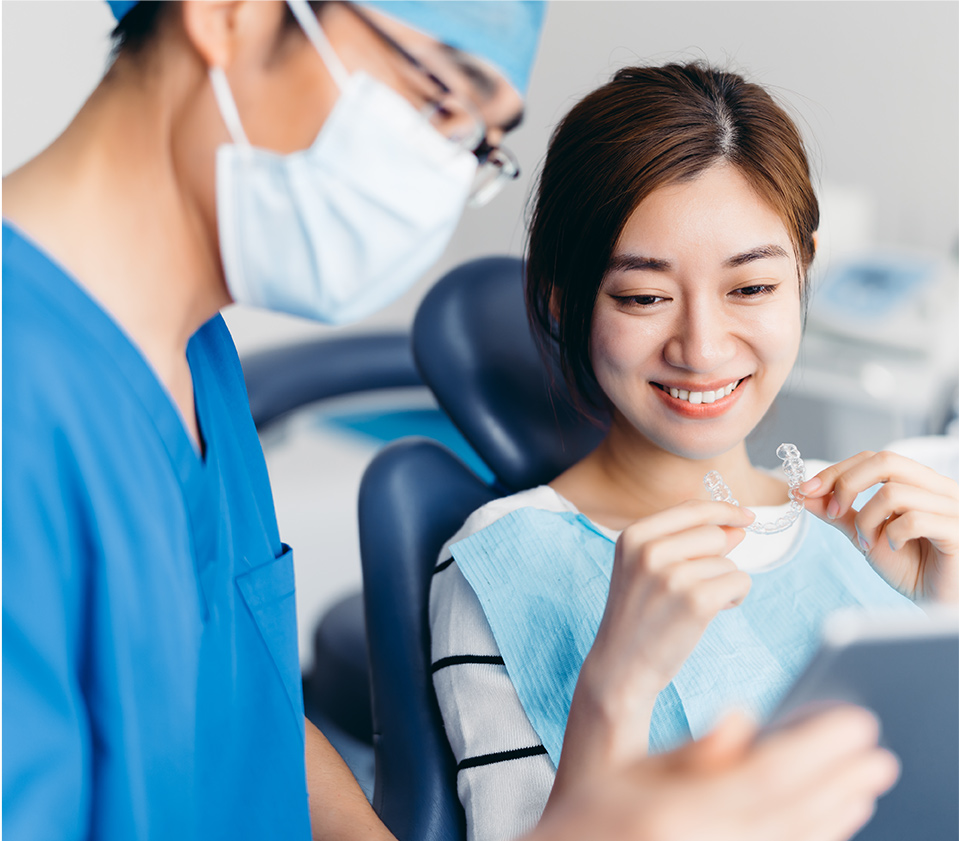 A dentist in blue scrubs and mask standing beside a smiling female patient holding clear orthodontic aligners